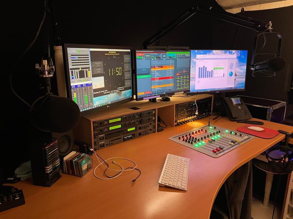 Radio control room with three monitors, mixing desk, and OnAirScreen clock with meters on the left screen