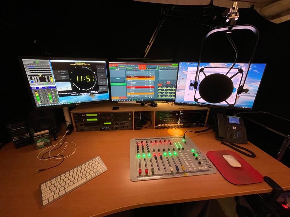 Radio control room with Airlite mixer, broadcast microphone, and OnAirScreen on the studio monitors